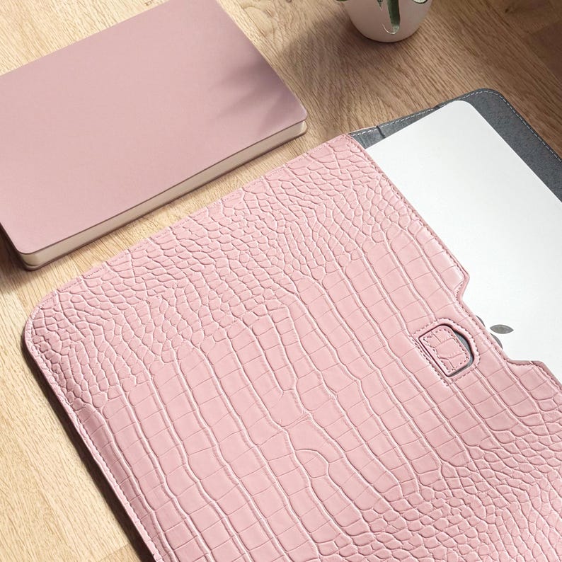 Op de afbeelding: Een roze laptoptas met een krokodillenleertextuur is open en onthult een laptop. Een bijpassend roze notitieboekje ligt naast de hoes. De items liggen op een houten oppervlak. De hoes heeft een D-ringsluiting.