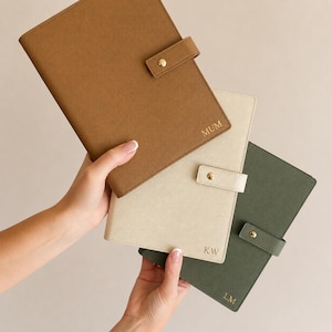 Op de afbeelding: Drie rechthoekige planners met gestructureerde covers in bruin, crème en olijfgroen. Elke planner heeft een goudkleurige drukknoopsluiting en goudkleurige letters in de rechterbenedenhoek met de tekst MUM, KW en LM respectievelijk.