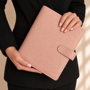 May include: A dusty pink, textured leather-like planner cover featuring a secure snap-button closure and the gold-embossed initials H.F. at the bottom right corner. The rectangular organizer has a pebbled surface and a clean, structured design.