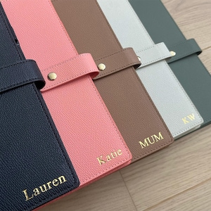 Op de afbeelding: Vijf rechthoekige planners met gestructureerde covers in marineblauw, roze, bruin, lichtgrijs en donkergroen. Elke planner heeft een gouden drukknoopsluiting en een gouden foliebedrukking met de namen Lauren, Katie, MUM, KW en LM.
