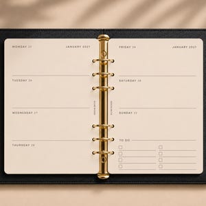 May include: An open black leather planner with cream-coloured pages and a gold-toned six-ring metal binder mechanism. The pages display a weekly layout for January 2027, featuring days of the week, dates, and a to-do list section with checkboxes.