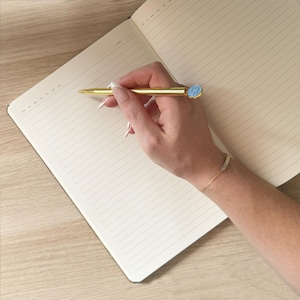K&ouml;nnte beinhalten: Ein goldener Stift mit hellblauem Akzent wird &uuml;ber einem offenen Notizbuch mit linierten Seiten gehalten. Der Stift ist so positioniert, als w&uuml;rde er auf die Seite schreiben. Ein goldenes Armband ist sichtbar.