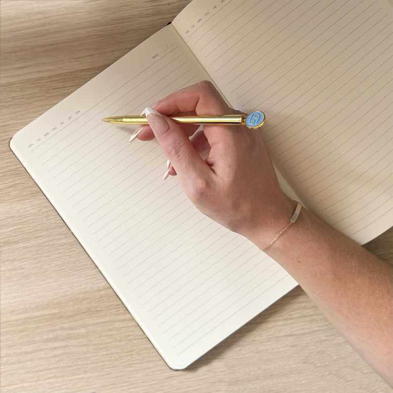 K&ouml;nnte beinhalten: Ein goldener Stift mit hellblauem Akzent wird &uuml;ber einem offenen Notizbuch mit linierten Seiten gehalten. Der Stift ist so positioniert, als w&uuml;rde er auf die Seite schreiben. Ein goldenes Armband ist sichtbar.