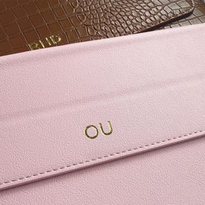May include: Close-up of two leather clutch bags. The top bag is brown with a crocodile texture and gold lettering that reads "PHD". The bottom bag is pink with gold lettering that reads "OU". Both bags have a stitched edge.