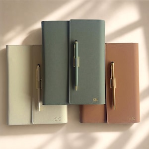 Op de afbeelding: Drie rechthoekige notitieboeken met gestructureerde covers in beige, saliegroen en lichtbruin. Elk notitieboek heeft een verticale penlus met een bijpassende balpen en is gepersonaliseerd met de gouden initialen G.C, S.R en T.K.