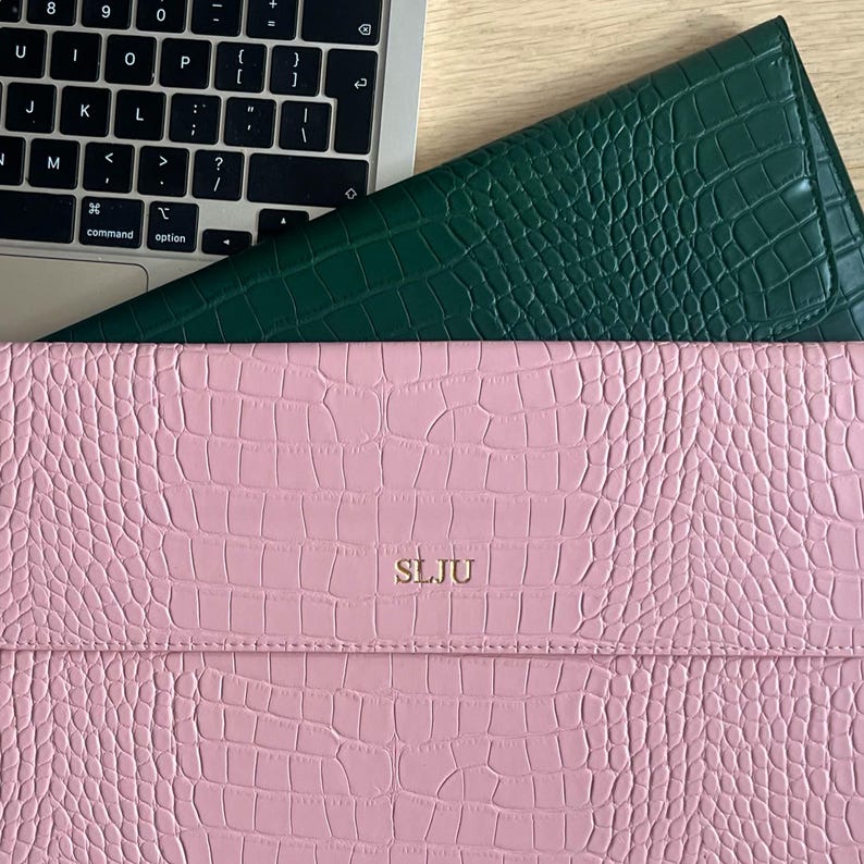 Op de afbeelding: Een roze en groene laptophoes van imitatieleer met een krokodillenleertextuur. De roze hoes heeft de initialen "SLJU" in goudreli&euml;f. Een laptoptoetsenbord is gedeeltelijk zichtbaar op de achtergrond.