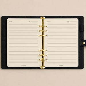 May include: A black leather-textured binder with a gold-toned six-ring metal mechanism. The interior features cream-colored lined paper with the printed text NOTES and Date at the top of each page. A black strap with a gold snap closure is attached.