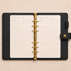 Op de afbeelding: Een zwarte planner met ledertextuur en een goudkleurig metalen ringbandmechanisme met zes ringen. De open planner toont twee crèmekleurige pagina's met de gedrukte tekst MEETING NOTES, Date, Start time, End time, Subject, Attendees, Notes en Action.