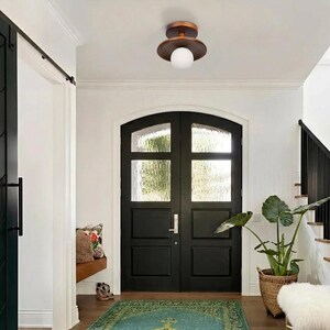 May include: A modern ceiling light fixture with a bronze finish and a white globe light. The light is mounted on a white ceiling in a hallway with black double doors and a green rug.