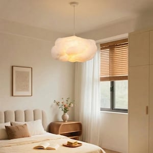 May include: A cloud-shaped pendant light fixture hangs from the ceiling in a bedroom. The light is white and has a soft, diffused glow. The bedroom has a neutral color palette with a bed, nightstand, and window with blinds.