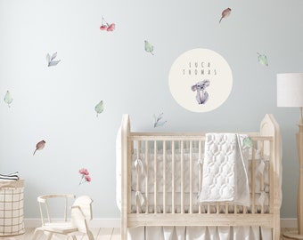 May include: A nursery scene with a light blue wall adorned with watercolor leaf and flower decals. A crib with a white quilt and a rocking horse are in the foreground. A circular decal features a koala and the name "LUCA THOMAS".