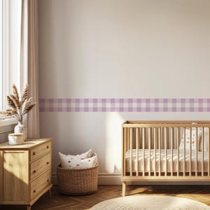 May include: A nursery interior featuring a wooden cot, chest of drawers, and a wicker basket. The wall has a light purple and white gingham border. Dried flowers in a vase sit on the chest, and a circular rug is on the floor.
