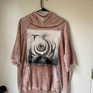 May include: A short-sleeved hooded sweatshirt in a faded, mottled terracotta color. The front features a large, rectangular patch with a black and white graphic illustration of a central eye surrounded by dark, claw-like shapes and a bird.