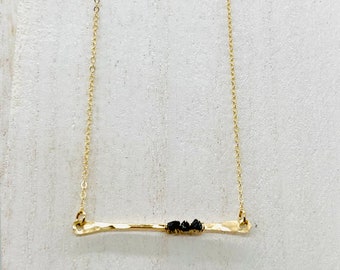 May include: A gold chain necklace with a thin, hammered gold bar pendant accented with black stones.