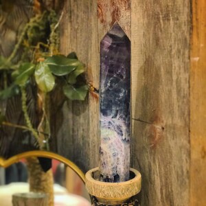 Könnte beinhalten: Eine violette und weiße Kristallspitze mit einer spitzen Spitze, die in einem Holzhalter sitzt. Der Kristall ist ein Naturstein mit einem einzigartigen Muster.