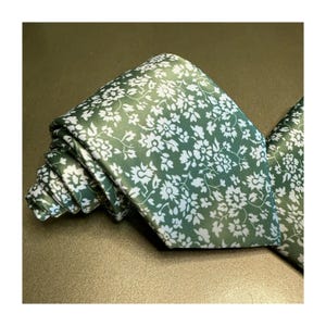 Op de afbeelding: Een opgerolde groene stropdas en bijpassende pochet. De stropdas en pochet hebben een herhalend patroon van kleine witte bloemmotieven. De stropdas is gemaakt van een gladde stof en is geschikt voor formele gelegenheden.