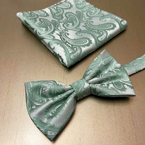May include: A green and white paisley patterned bow tie and pocket square set. The bow tie is tied and the pocket square is folded.