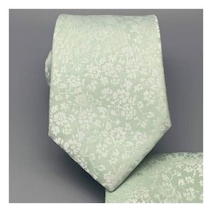 Könnte beinhalten: Eine hellgrüne gemusterte Krawatte mit weißem Blumenmuster. Die Krawatte ist aus seidenartigem Material und hat eine spitze Spitze.