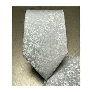 May include: A light gray tie and matching pocket square set. The tie and pocket square feature a repeating floral pattern in a lighter shade of gray. The tie is a standard width and is made of a smooth fabric.