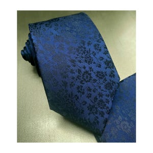 May include: A navy blue tie with a repeating floral pattern in a darker shade of blue. The tie is folded and appears to be made of a textured fabric. The floral design adds a touch of elegance.