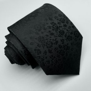 May include: A black silk tie with a subtle floral pattern. The tie is rolled up and ready to be worn.