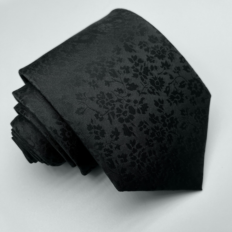 May include: A black silk tie with a subtle floral pattern. The tie is rolled up and ready to be worn.