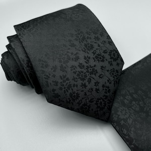 May include: A black silk tie with a subtle floral pattern. The tie is rolled up and the pattern is visible on the front and back.