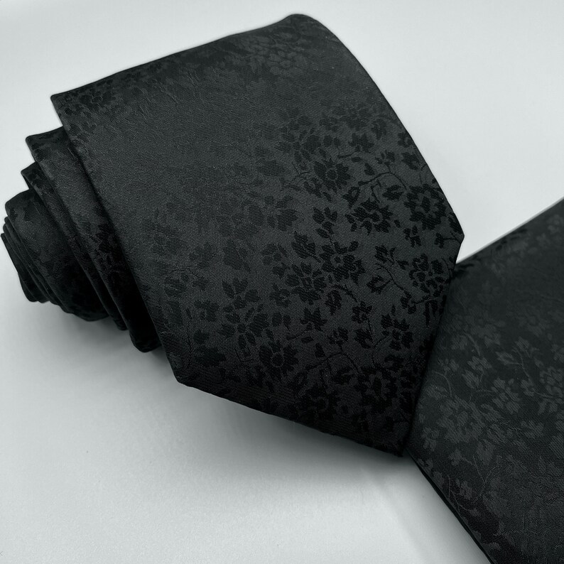 May include: A black silk tie with a subtle floral pattern. The tie is rolled up and the pattern is visible on the front and back.