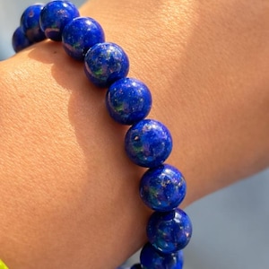 May include: A bracelet featuring large, round, deep blue beads with flecks of gold and green. The beads are strung together, creating a bold and eye-catching accessory. The bracelet is shown on a wrist.