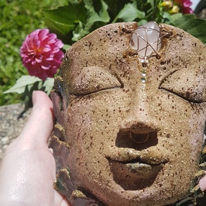 May include: A handmade ceramic face planter with closed eyes, a small nose ring, and a rose quartz crystal. The planter is a light brown colour with a textured surface. The background includes pink dahlias and green foliage.
