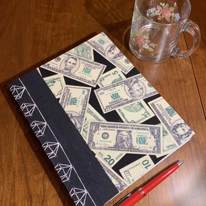 Puede incluir: Un cuaderno negro con un diseño de puntadas de diamante blanco en el lomo. La portada está decorada con un collage de billetes de dólar estadounidense. Un bolígrafo rojo está sobre una superficie de madera junto al cuaderno.