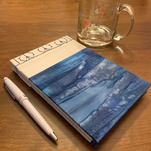 Puede incluir: Un cuaderno azul y blanco con una cubierta decorativa y un bolígrafo blanco sobre una mesa de madera.