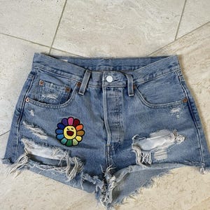 Puede incluir: Shorts vaqueros azul claro con detalles desgastados y un parche de flores de colores. Los shorts tienen cierre de botón, bolsillos delanteros y bordes deshilachados. El parche presenta un diseño de flor sonriente con colores arcoíris.