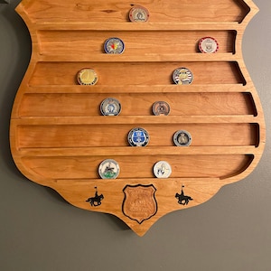 Op de afbeelding: Een houten display in de vorm van een politiebadge met zeven planken om challenge-munten op te bergen. De display heeft aan elke kant een zwart silhouet van een politieagent te paard en in het midden de woorden "Police" en "Machete Group".