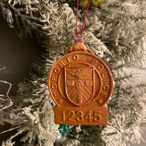 Könnte beinhalten: Ein hölzerner Weihnachtsbaumschmuck in Form eines Abzeichens der Toronto Police mit der Nummer 12345 auf der Unterseite.