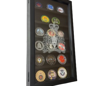 May include: A black wooden shadow box display case with a glass front, showcasing a collection of challenge coins. The coins feature various designs, including military insignia, emblems, and logos. The shadow box is designed to protect and display the coins.