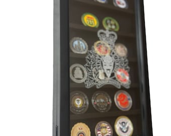 Op de afbeelding: Een zwarte houten schaduwdoos display case met een glazen voorzijde, die een verzameling challenge-munten toont. De munten hebben verschillende ontwerpen, waaronder militaire insignes, emblemen en logo's. De schaduwdoos is ontworpen om de munten te beschermen en te tonen.