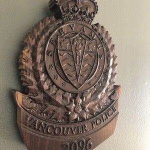 Puede incluir: Una placa de madera con una imagen tallada del escudo de armas del Departamento de Policía de Vancouver. El escudo presenta una corona, un escudo con un patrón de ondas y las palabras "Servir y proteger". La placa también incluye las palabras "Policía de Vancouver" y el número "2096".