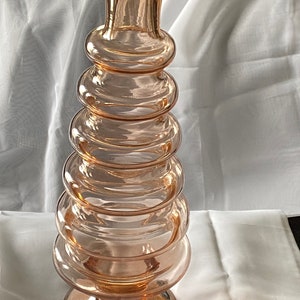 May include: A peach-colored glass vase with a stacked, circular design. The vase has a wide base and a narrow top, with a series of rounded rings creating its unique shape. The glass has a smooth, glossy finish.