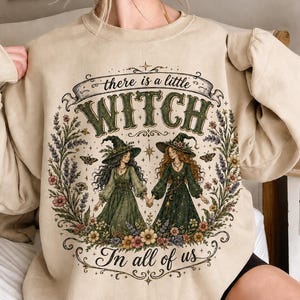 May include: A beige crewneck sweatshirt featuring a graphic print of two figures in witch hats and long dresses holding hands. The design includes floral borders, stars, and the text "there is a little WITCH In all of us" in a decorative font.