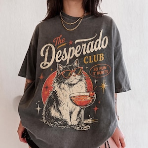 May include: A charcoal gray t-shirt featuring a graphic of a cat wearing red sunglasses next to a cocktail glass. The text reads The Desperado CLUB and SO FUN IT HURTS, surrounded by star accents and a red circular background element.