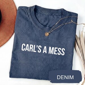 May include: A navy blue t-shirt with the text "CARL'S A MESS" in white. A gold-colored necklace with a pendant is draped across the shirt. The word "DENIM" is printed on a navy blue banner.