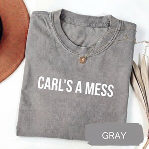 May include: Gray crew neck t-shirt with the text "CARL'S A MESS" in white block letters. A gold pendant necklace rests on the shirt. The word "GRAY" is written on a gray paint swatch.