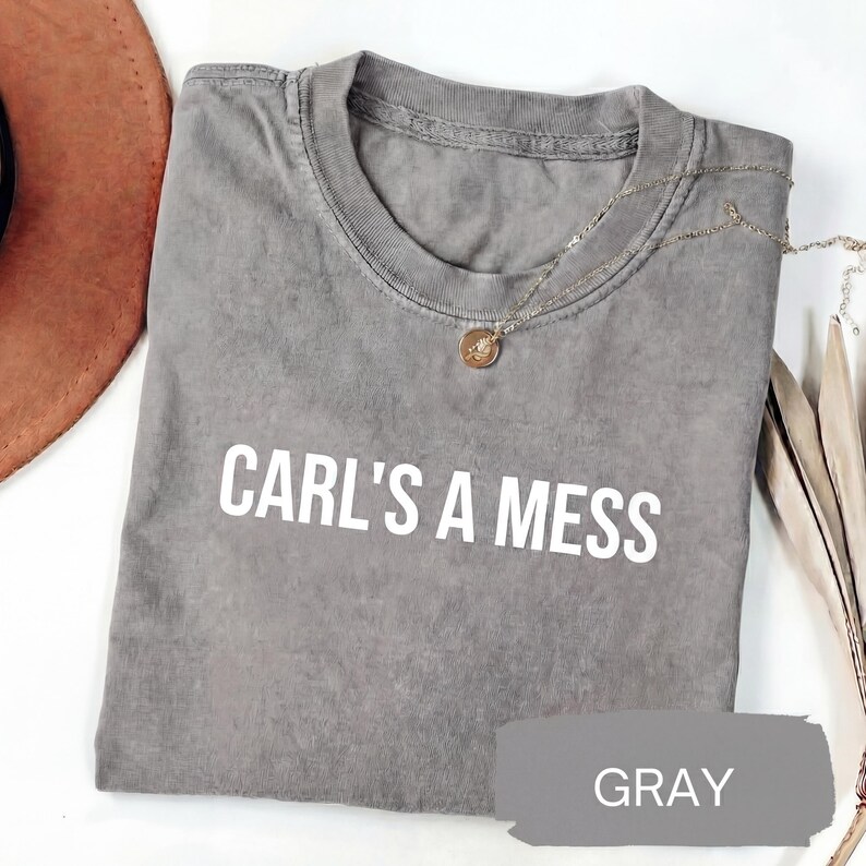 May include: Gray crew neck t-shirt with the text "CARL'S A MESS" in white block letters. A gold pendant necklace rests on the shirt. The word "GRAY" is written on a gray paint swatch.