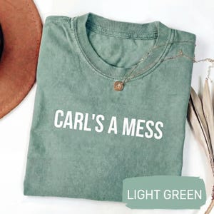 May include: Light green t-shirt with the text "CARL'S A MESS" in white. The shirt has a crew neck and short sleeves. A gold-colored necklace with a pendant is draped on the shirt. The words "LIGHT GREEN" are in a green rectangle.
