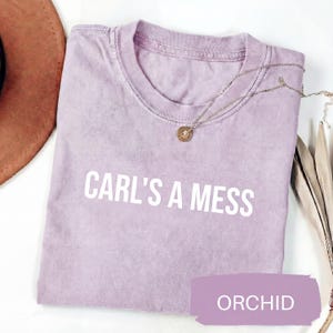 May include: Orchid-colored t-shirt with the text "CARL'S A MESS" in white. A gold necklace with a pendant is draped across the shirt. The shirt is folded and has a vintage, washed look.