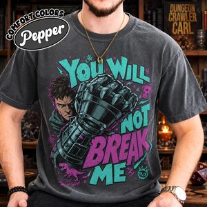 Pode incluir: Uma t-shirt cinzento-carvão de manga curta com uma impressão gráfica de uma pessoa e uma grande manopla blindada. O texto "YOU WILL NOT BREAK ME" está em turquesa e roxo. O design inclui um pequeno dinossauro cor-de-rosa.