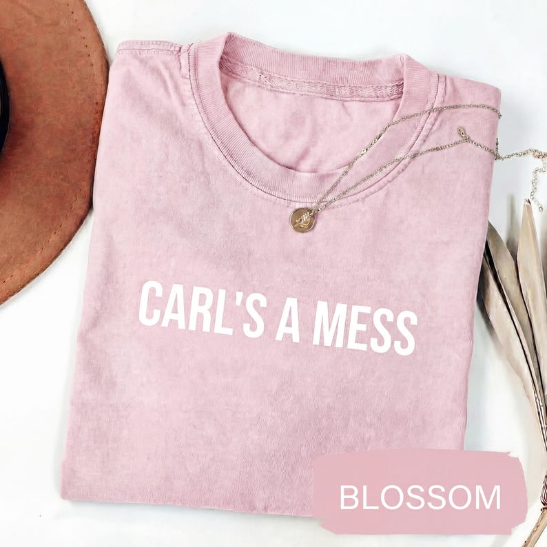 May include: A light pink t-shirt with the text "CARL'S A MESS" in white block letters. A silver necklace with a small pendant is draped across the shirt. The word "BLOSSOM" is printed on a pink rectangle.