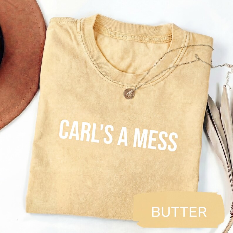 May include: Butter-colored t-shirt with the text "CARL'S A MESS" in white block letters. A silver necklace with a small pendant rests on the shirt. The shirt is folded and has a slightly faded appearance.