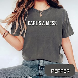 May include: A dark gray t-shirt with the text "CARL'S A MESS" in white block letters. The shirt is a crew neck style and appears to be made of a soft, comfortable material. The word "PEPPER" is displayed in a dark gray rectangle at the bottom of the image.